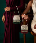 Two women seated beside a wooden chair, each holding a 925 silver handbag with pearl bead straps and dangling beaded semi precious stones fringe. One bag has green beads at the bottom and the other has red beads, against a dark teal studio background.