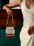 A woman in a dress holds a 925 silver bag with a pearl and red bead strap. The bag features intricate silver nakaashi, and red and white semi precious stones tassels, set against a dark wooden cabinet and teal wall background.